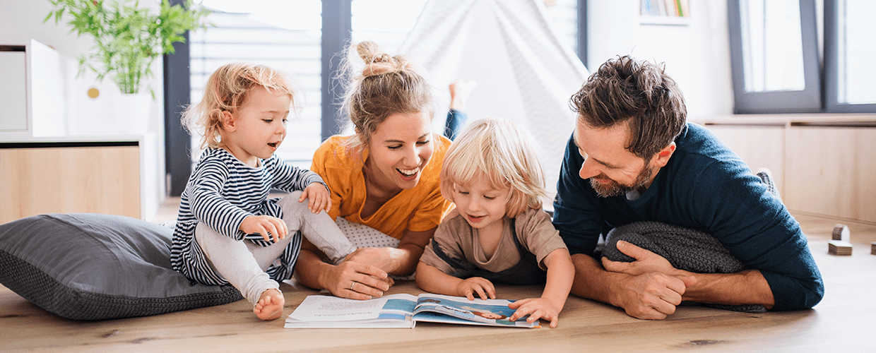 Eine junge Familie liegt auf dem Boden des Wohnzimmers. Zwei blonde Kinder sowie Mutter und Vater schauen gemeinsam in ein aufgeschlagenes Buch